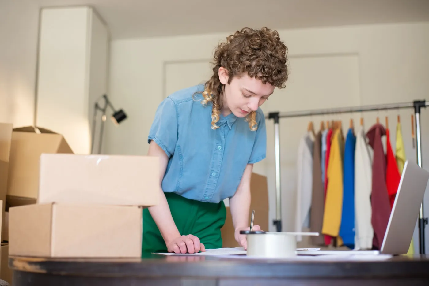 A clothing shop owner noting orders beside packed boxes and a garment rail