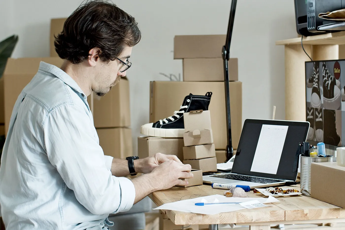 An online shoe seller boxing orders at a desk beside a laptop and stacked cartons